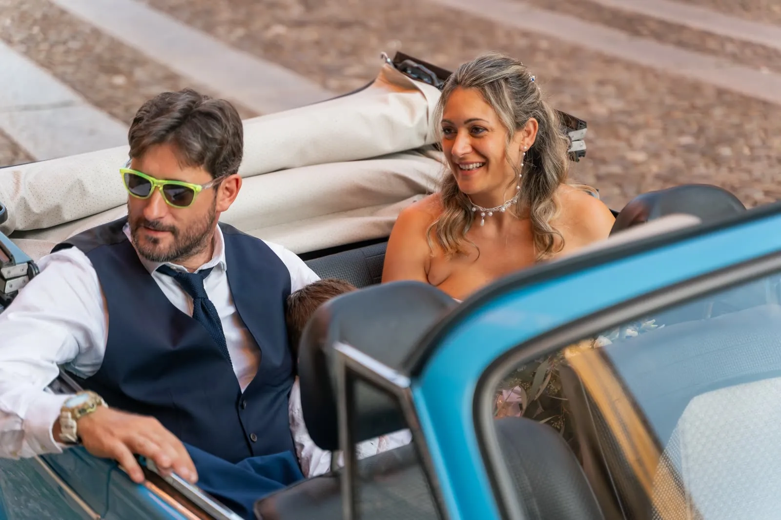 Gli sposi nel Maggiolone cabriolet azzurro con la capote abbassata, in piazza: lui con gli occhiali da sole giallo fluo, lei che sorride