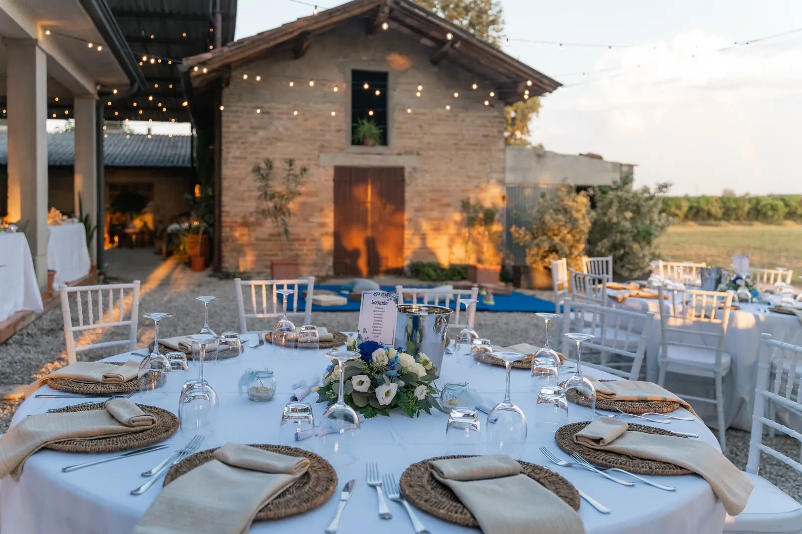 Un tavolo rotondo apparecchiato all'aperto con piatti di paglia e fiori blu e bianchi, davanti a un casolare di mattoni illuminato dal tramonto