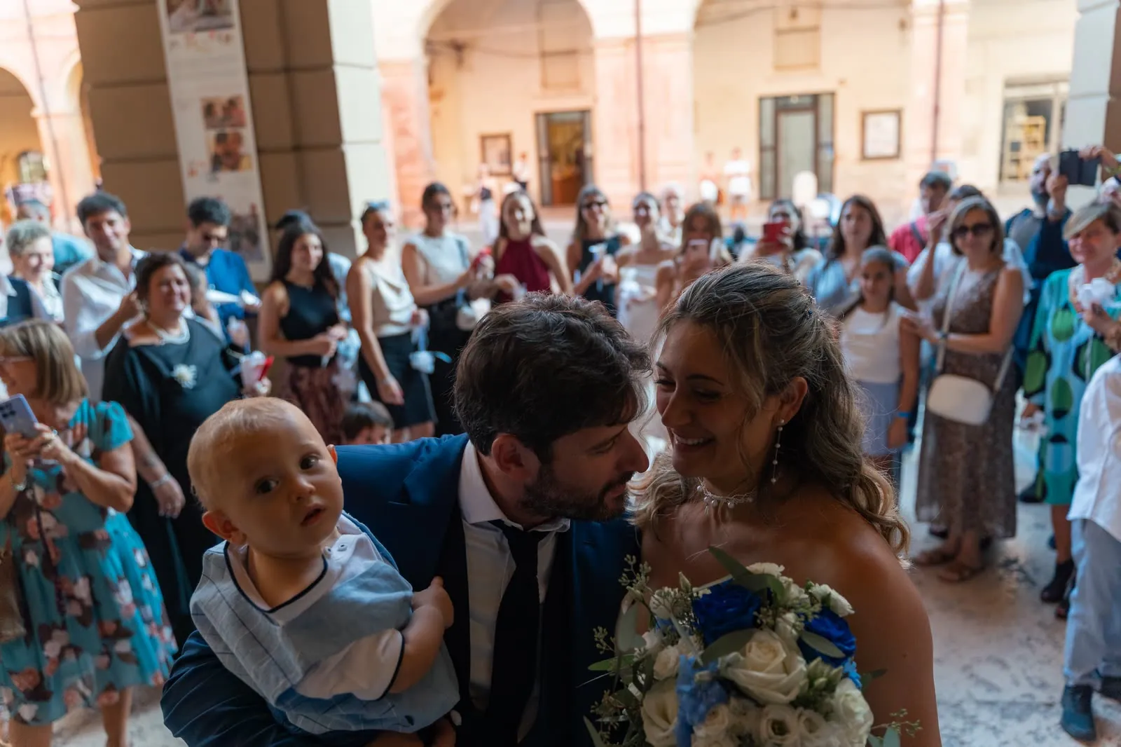 Gli sposi accolti sotto i portici dagli invitati: lui si china verso di lei, sorridente, con un bambino in braccio