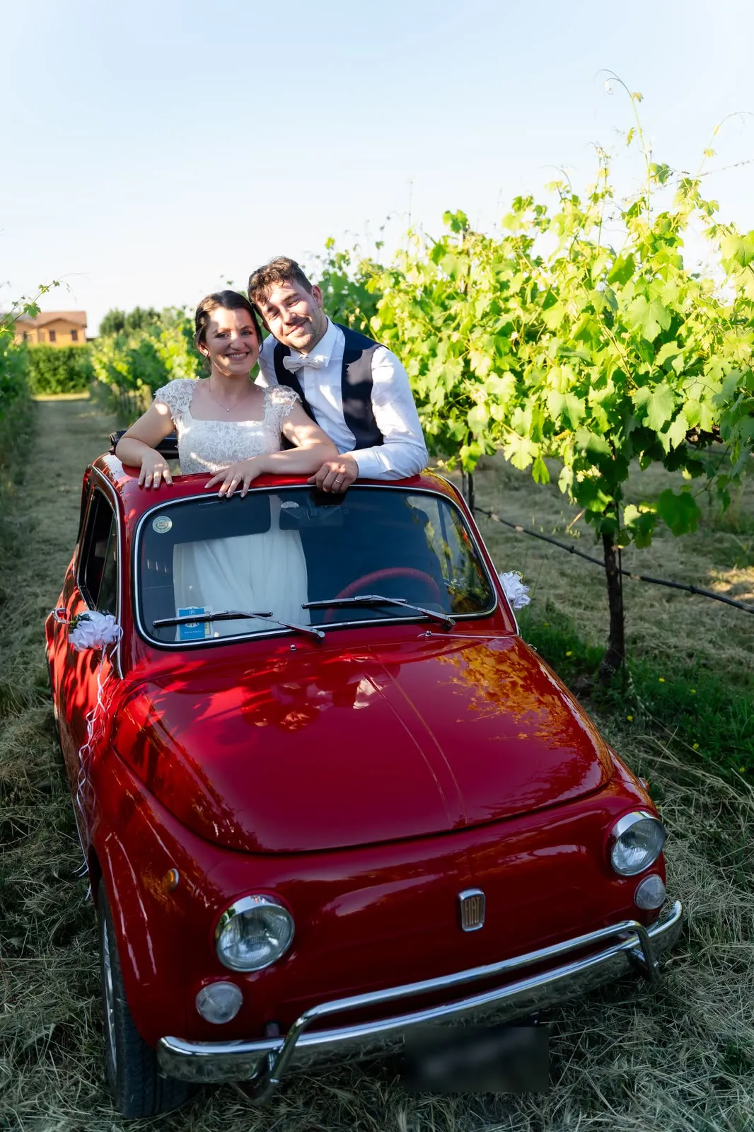 Gli sposi sorridono affacciati dal tettuccio aperto della Fiat 500 rossa, tra i filari