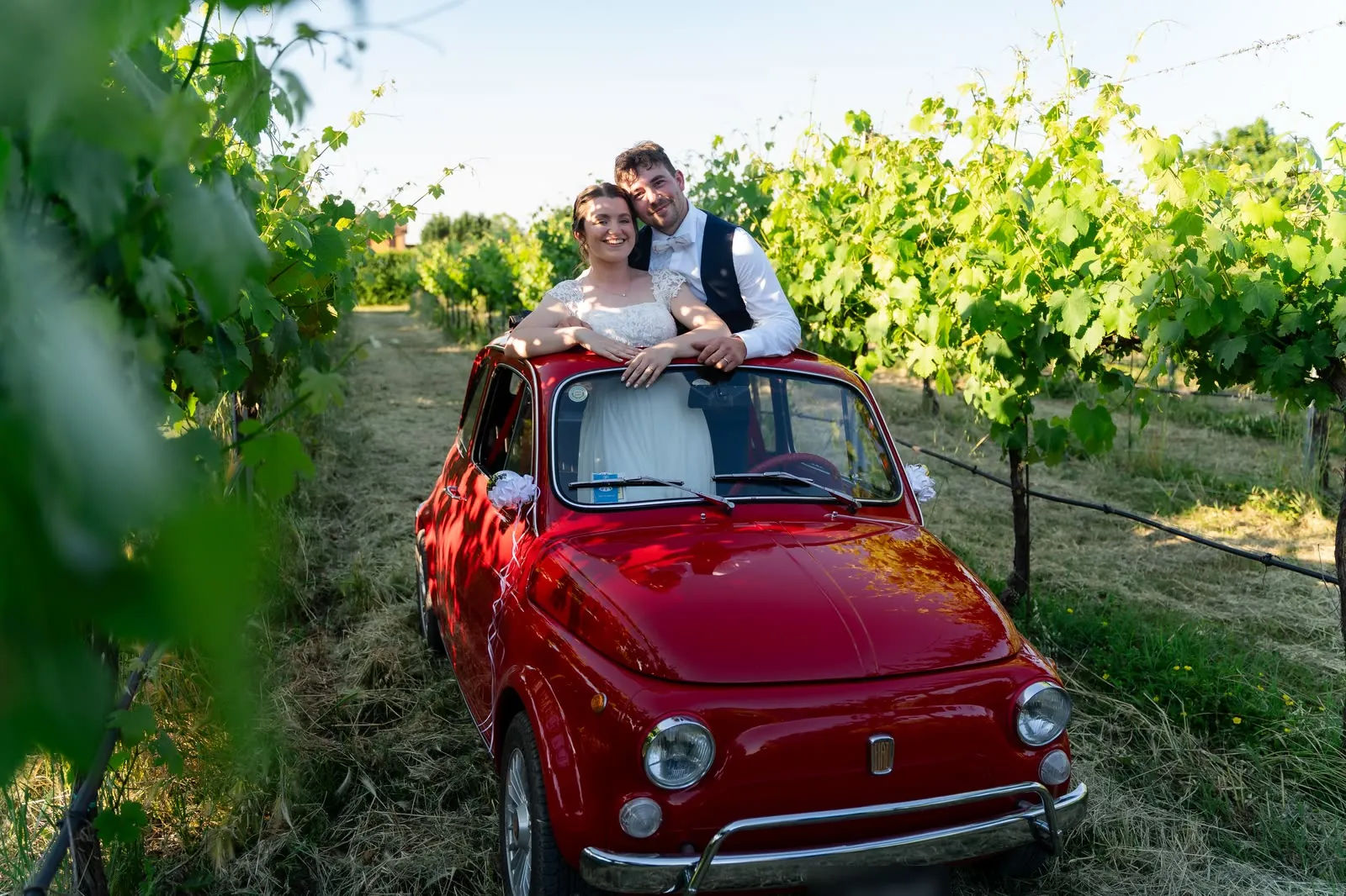 Gli sposi si affacciano sorridenti dal tettuccio aperto della Fiat 500 d'epoca, in mezzo ai filari