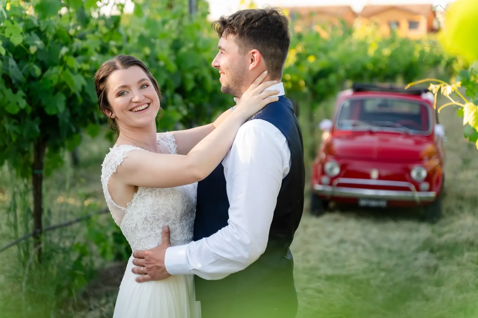 La sposa ride abbracciando lo sposo tra i filari, con la Fiat 500 rossa sfocata alle spalle