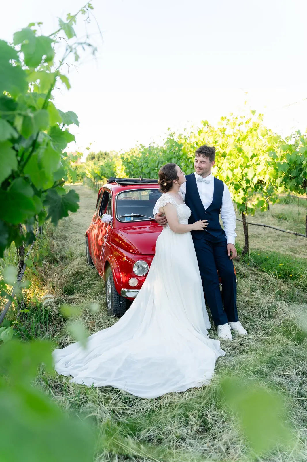 Gli sposi abbracciati accanto alla Fiat 500 rossa, con lo strascico dell'abito disteso sull'erba tra i filari
