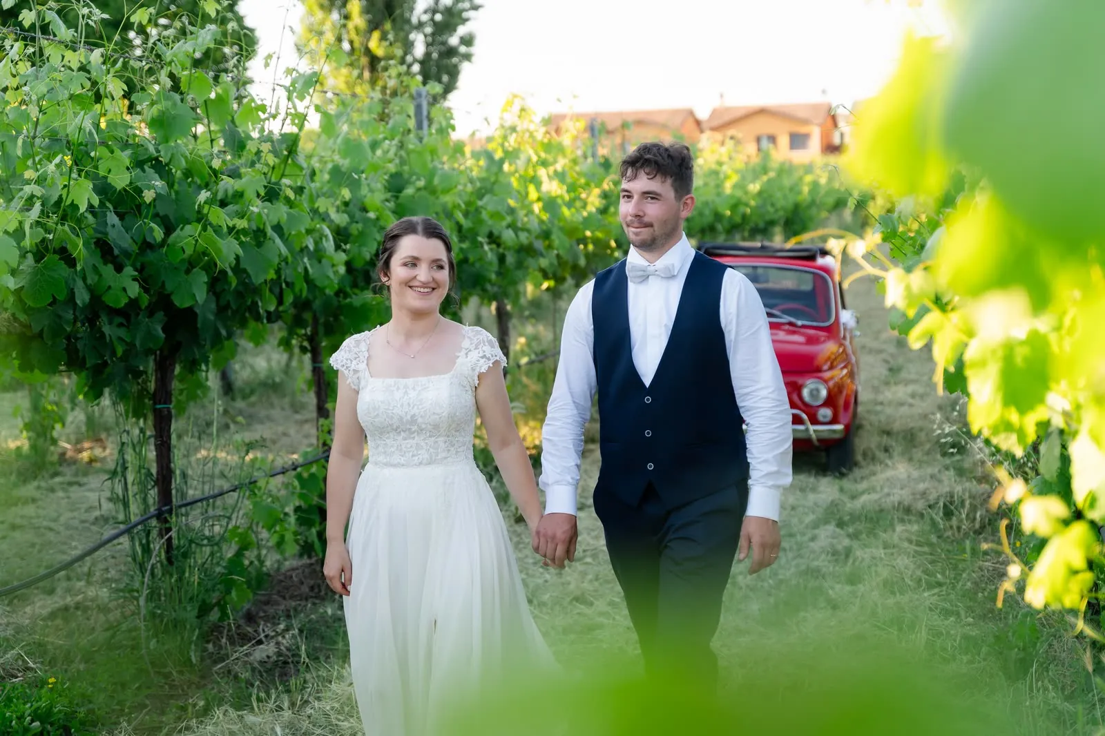 Gli sposi passeggiano mano nella mano tra i filari della vigna, con la 500 rossa alle spalle