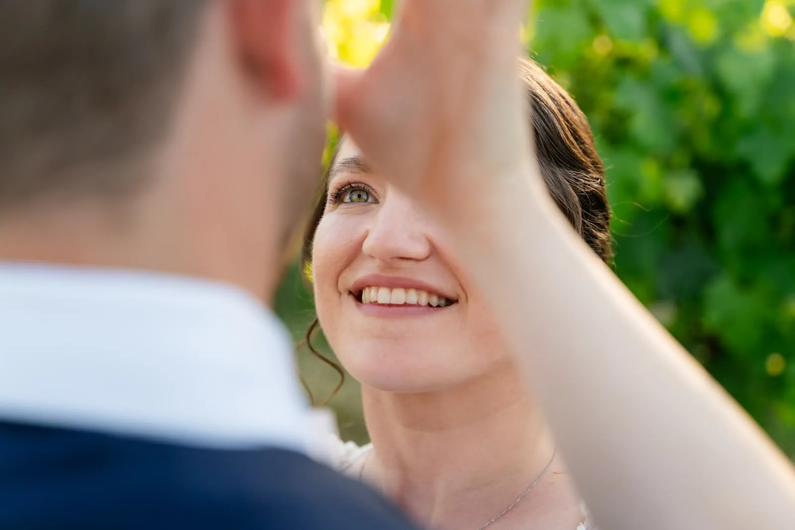 Primo piano del sorriso della sposa, con la mano dello sposo vicino al viso, tra le foglie della vigna