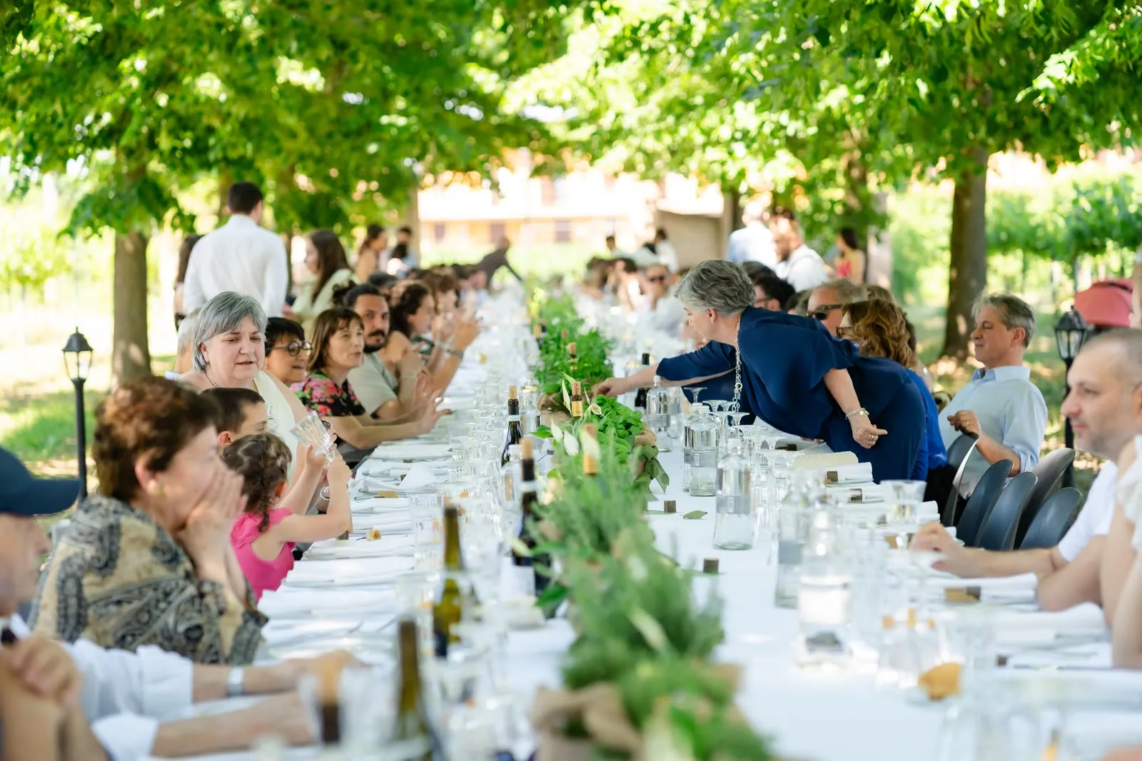 Una lunga tavolata all'aperto sotto gli alberi, con gli invitati seduti uno di fronte all'altro