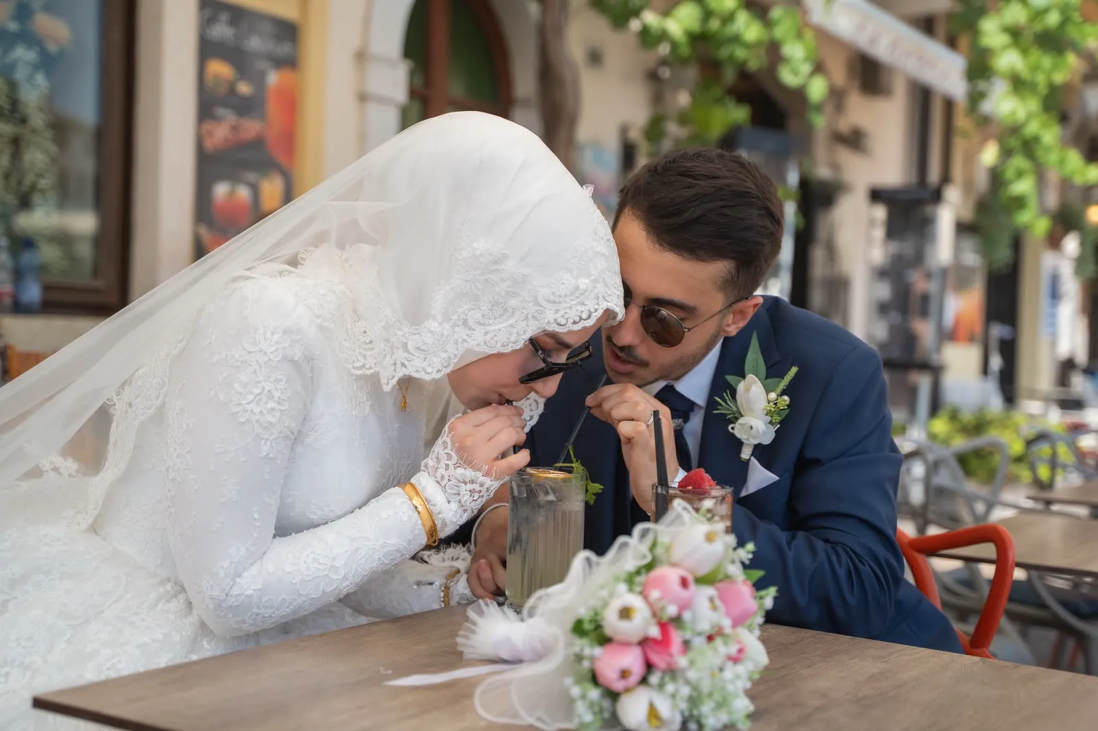 La sposa beve da una cannuccia al tavolino di un bar mentre lo sposo, con gli occhiali da sole, le sussurra qualcosa