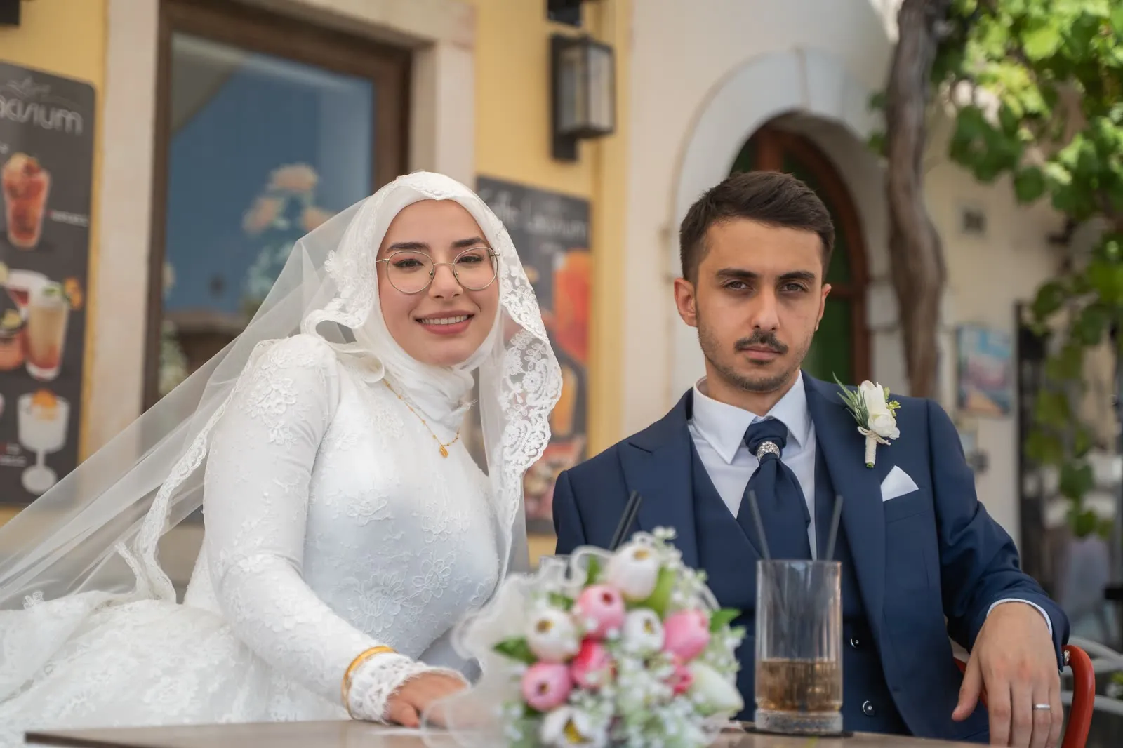 La sposa sorride all'obiettivo mentre lo sposo posa serissimo, seduti al tavolino di un bar con il bouquet in primo piano