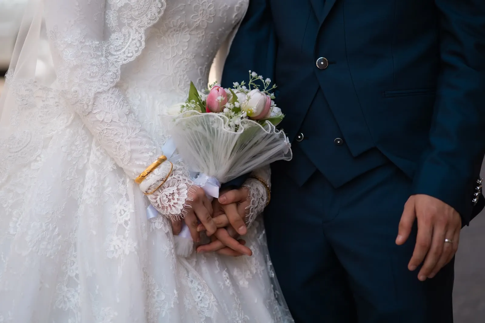 Le mani intrecciate degli sposi, con i polsini di pizzo, i bracciali dorati e il bouquet di tulipani rosa e bianchi