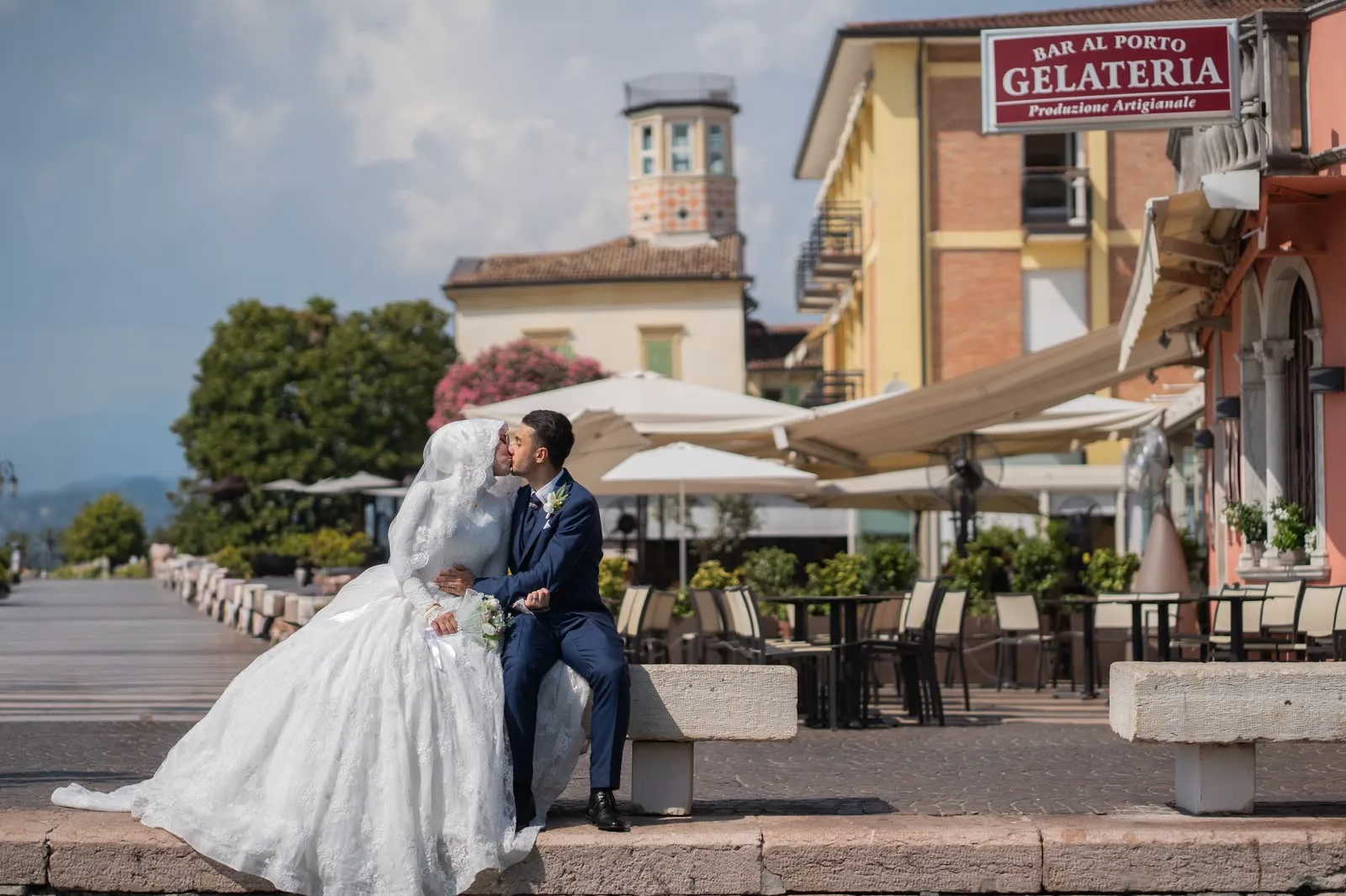 Gli sposi si baciano seduti su una panca di pietra sul lungolago di Lazise, con i palazzi color pastello e una torretta alle spalle