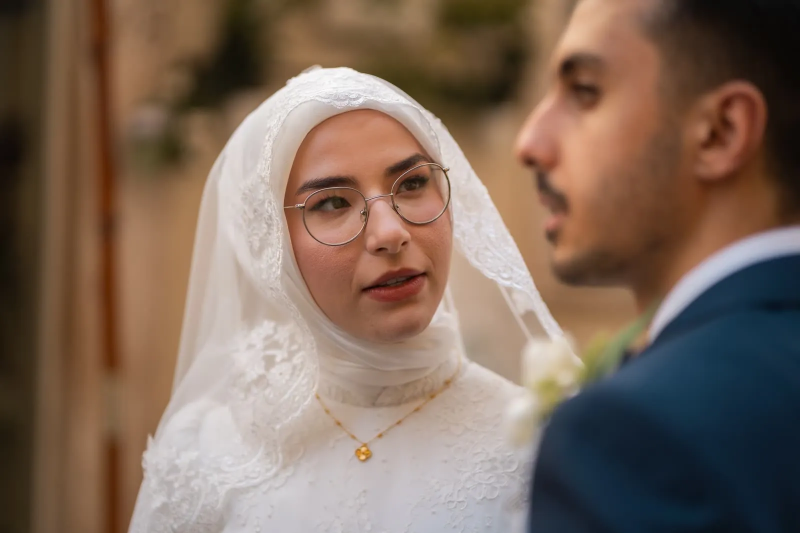 Primo piano della sposa con il velo di pizzo e gli occhiali, che guarda lo sposo sfocato in primo piano
