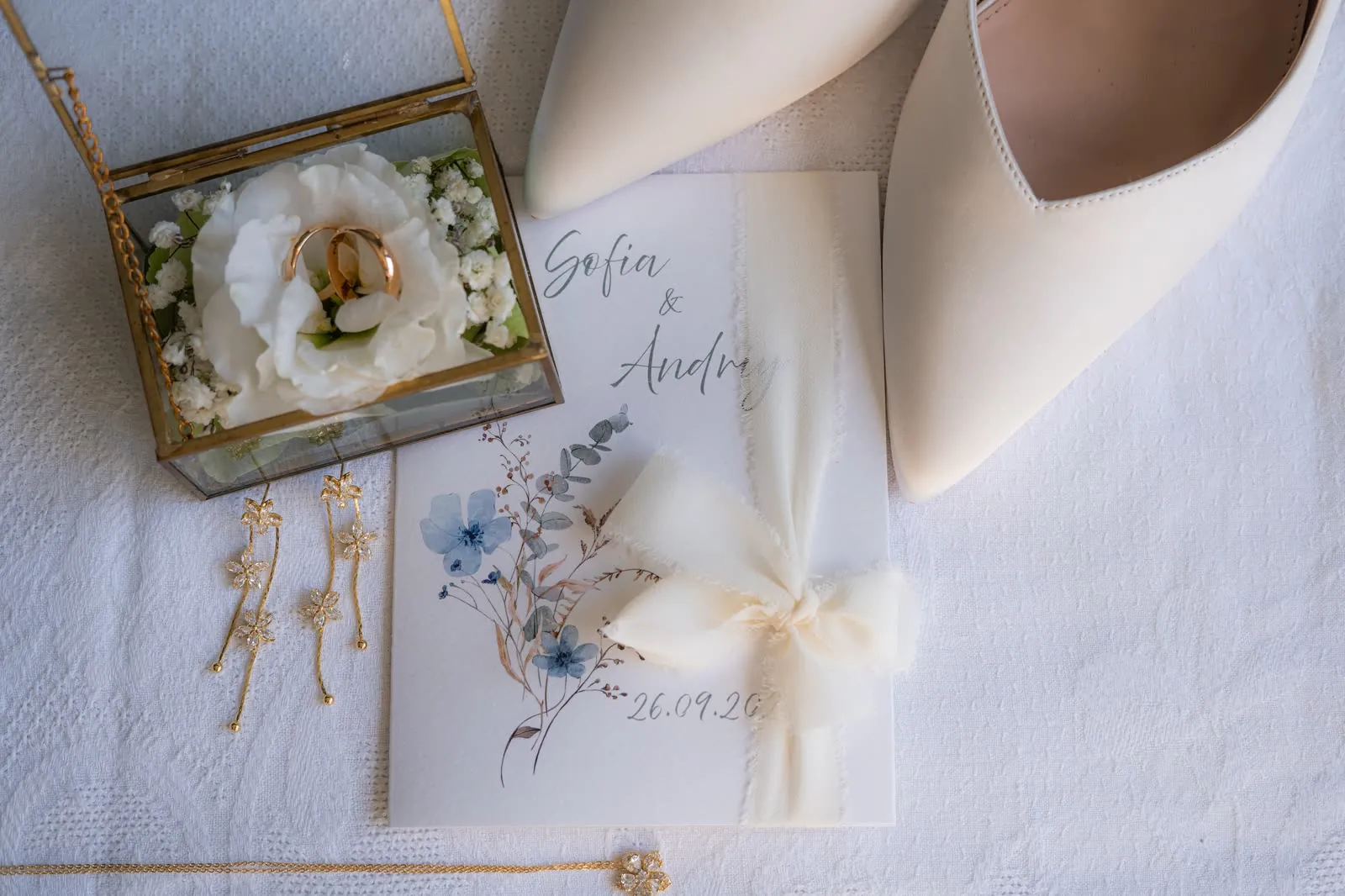 Le fedi in una scatola di vetro con un fiore bianco, accanto alle scarpe, agli orecchini e all'invito con i nomi di Sofia e Andriy