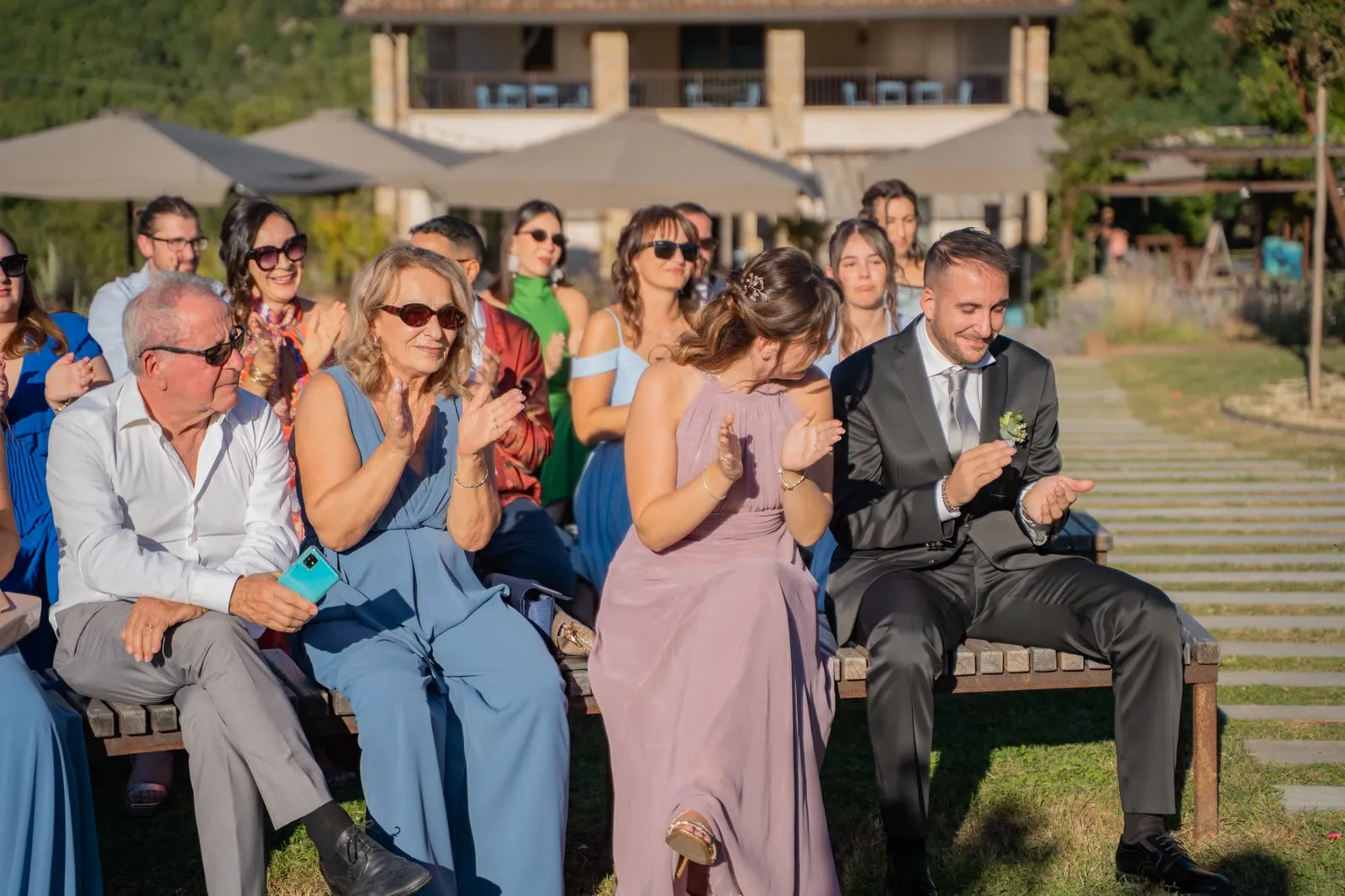 Gli invitati applaudono seduti sulle panche di legno durante la cerimonia all'aperto