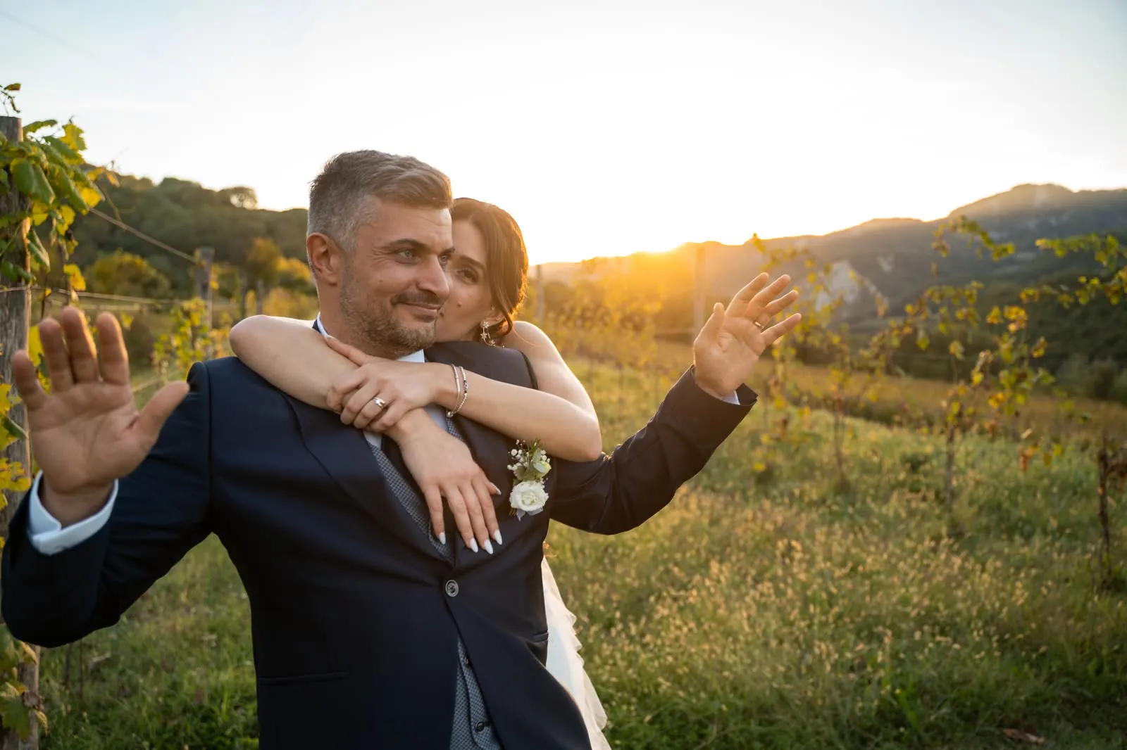 Lo sposo alza le mani in segno di resa mentre la sposa lo abbraccia da dietro e gli dà un bacio, tra le vigne al tramonto